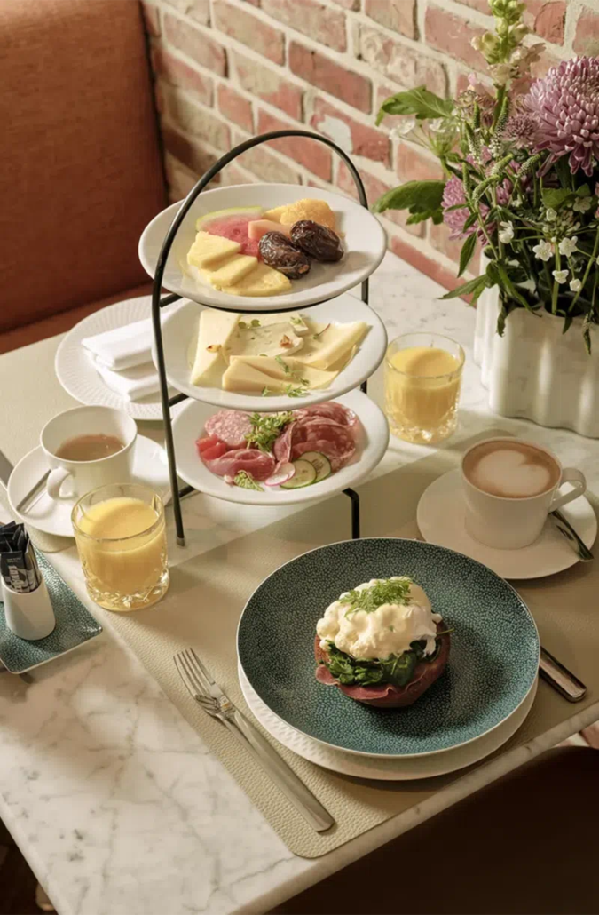 Gedeckter Frühstückstisch mit Kaffeespezialitäten, Orangensaft, einer frischen Eierspeise sowie einer Etagere in der Mitte des Tisches mit Wurst, Käse und Obst